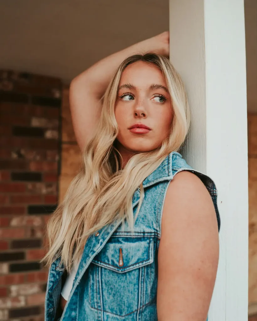 Person with long blonde wavy hair leaning against a white door frame with arm raised above head. Wearing a sleeveless blue denim vest over a dark top. Warm indoor lighting creates contrast with exposed brick wall background, giving the portrait a rustic, moody atmosphere.
