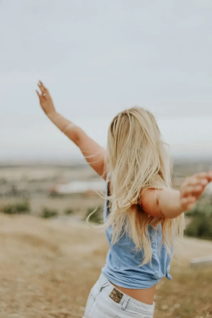 Back view of a person with blonde hair styled in a braided half-updo, sitting in a white wooden chair outdoors. They wear a light blue striped off-shoulder top and denim shorts against a blurred green and golden landscape background.